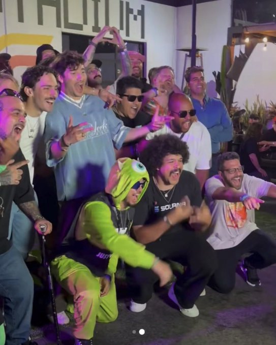 A large group of men pose energetically together at an outdoor nighttime party, with one person wearing a bright green Mike Wazowski costume.