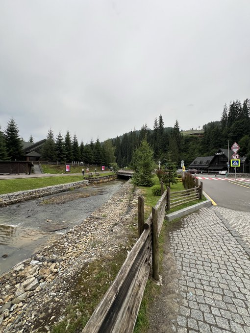 An overcast outdoor scene shows a shallow rocky stream flowing beside a wooden fence, with a cobblestone path, pine-covered hills, buildings, and a paved road visible in the background.