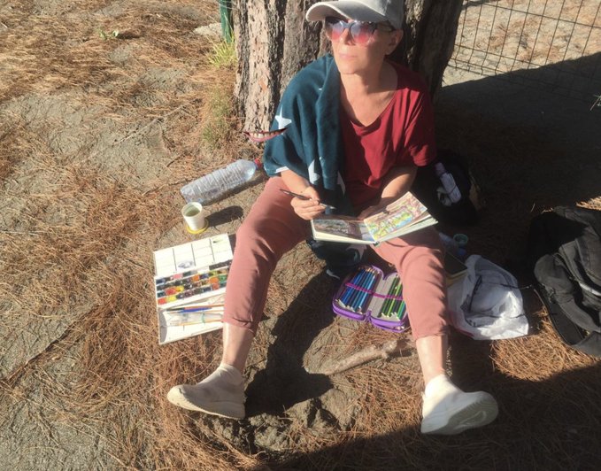 A person wearing a cap and sunglasses sitting against a tree on pine-needle covered ground, painting in a watercolor sketchbook with colored pencils and a water bottle nearby.