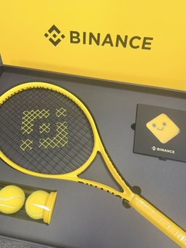 A promotional product image showing a yellow tennis racket branded with Binance logos, two yellow tennis balls, and a Binance card, all displayed in front of a yellow Binance-branded box.