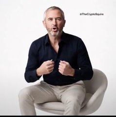 A man in a dark shirt and light pants sits in a chair with his fists raised, with the handle "@TheCryptoQuaire" visible in the corner.