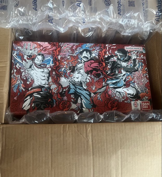 An opened cardboard shipping box containing an anime-themed Bandai Namco figure box featuring characters with red flame motifs, protected by air pillow packaging branded 'AIRplus'.