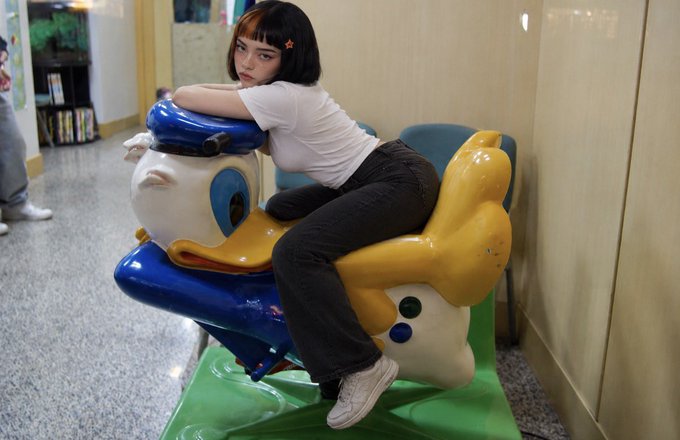 A young woman with a black bob haircut rests on a blue and yellow duck-shaped kiddie ride inside what appears to be a mall or arcade.