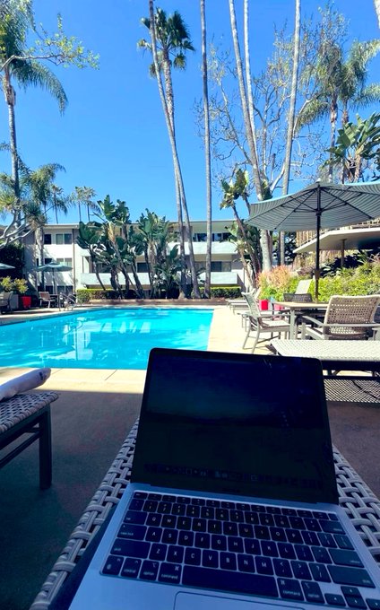 Outdoor workspace photo featuring a laptop on a table beside a swimming pool, with palm trees and a building in the background.