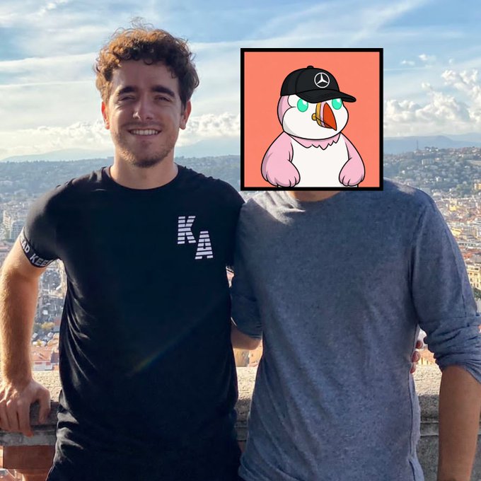 A man in a black t-shirt smiling next to a person whose face is obscured by an animated pink bird wearing a black Mercedes cap.