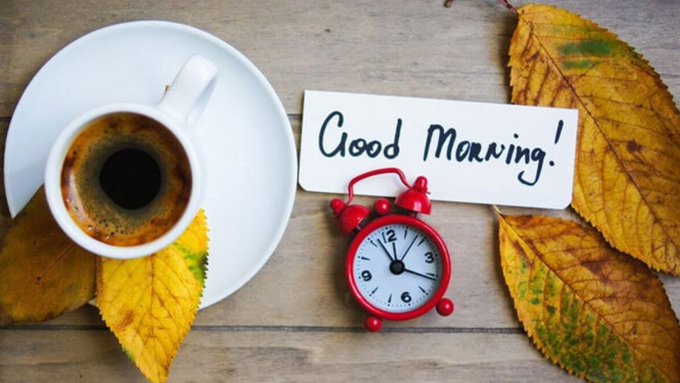 A flat-lay photograph showing a cup of black coffee, autumn leaves, a small red alarm clock, and a handwritten note that reads "Good Morning!" on a wooden surface.