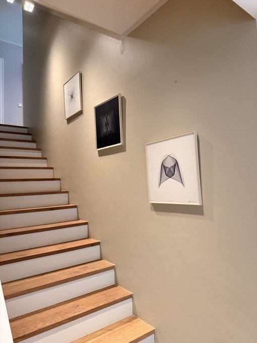 A wooden staircase ascends alongside a beige wall displaying three framed black-and-white artworks—including a geometric fox design—appearing to be the NFT pieces referenced by @benstraussphoto being showcased in a home setting.