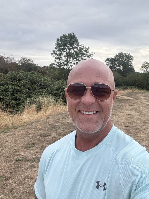 A selfie of a bald man with a gray beard wearing aviator sunglasses and a light blue Under Armour t-shirt, standing in a grassy field with trees and a cloudy sky in the background.