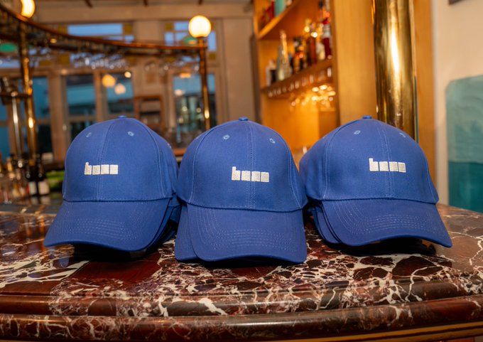 Three blue baseball caps with white embroidered text sit on a marble countertop inside what appears to be a bar, with bottles and warm lighting in the background.