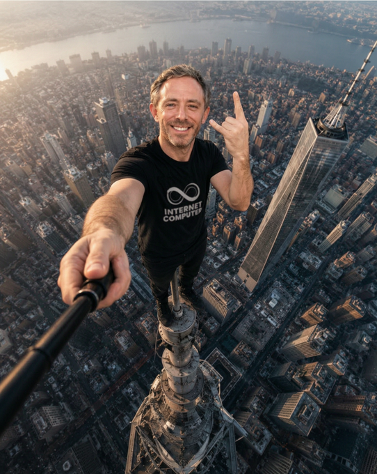 A man wearing a black 'Internet Computer' t-shirt takes a selfie with a selfie stick while standing atop a skyscraper antenna overlooking a city skyline.
