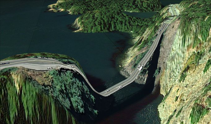 An aerial view of a winding two-lane road carved along the edge of steep, mossy green mountains, crossing a bridge over a dark body of water.