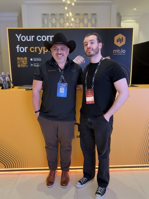 The image features two men standing next to each other, both wearing black shirts and hats. They appear to be posing for a photo or attending an event together. The background of the image shows a yellow wall with a sign that reads "Your corr". There is also a computer monitor on the wall.
