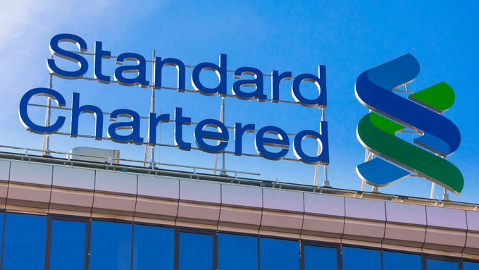 The image shows a large blue "Standard Chartered" sign mounted on the rooftop of a building against a clear blue sky, featuring the bank's distinctive blue and green logo. This relates directly to the post about Standard Chartered being added to the EU's MiCA register as an approved crypto service provider, highlighting the bank's expanding role in the regulated cryptocurrency sector.