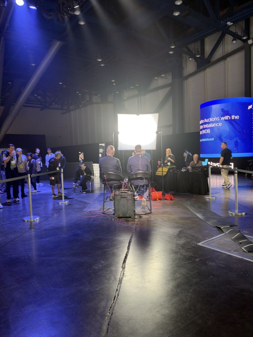 A dimly lit conference stage features a panel discussion with a few guests seated at a table, while a large screen displays text referencing "analytics with the [mak]esence" and "#NDSI" alongside what appears to be a CNBC segment number, suggesting the speaker briefly appeared on a CNBC broadcast.