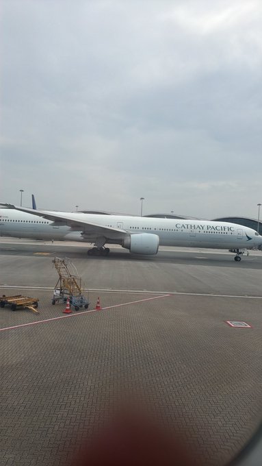 A Cathay Pacific airplane is parked on an airport tarmac under a cloudy sky, with traffic cones and ground equipment visible in the foreground.