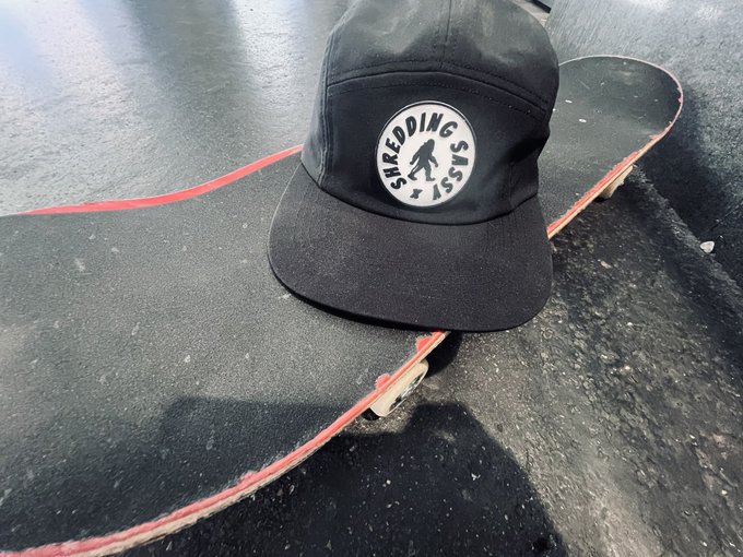 A black 'Shredding Sesh' camp cap resting on the grip tape of a skateboard.