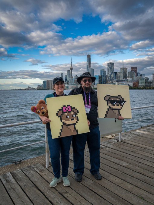 Two people stand on a wooden pier holding pixel art paintings of a character and a raccoon, with a city skyline visible across the water in the background.