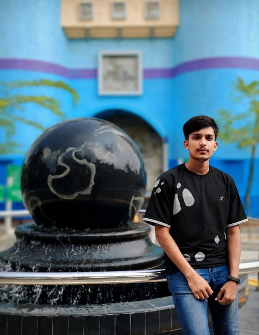 A young man in a black patterned t-shirt and jeans stands next to a large black marble sphere fountain in front of a bright blue building.