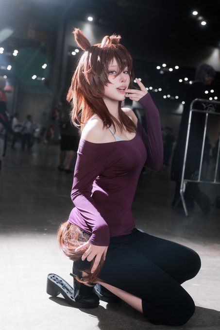 A cosplayer with animal ears, a brown wig, and a furry tail poses indoors while wearing a maroon off-the-shoulder top and black pants.
