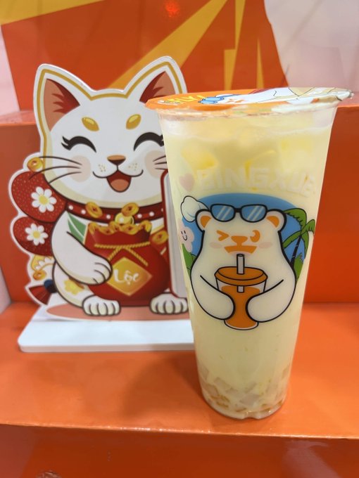 A photo of a clear plastic cup filled with a light-colored beverage, decorated with a cartoon bear wearing sunglasses and holding a coconut drink, sitting next to a smiling lucky cat (maneki-neko) display inside a store.