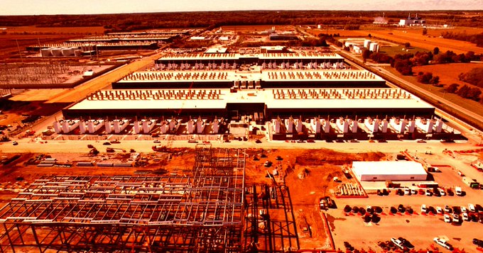 An aerial photograph of a massive data center complex under construction, with rows of building rooftops, exposed steel framework, parked vehicles, and surrounding rural landscape rendered in a heavy reddish-orange filter that suggests heat or overbuilding.

The photo of a half-constructed data center campus, tinted in harsh red-orange tones, visually reinforces the post's message about the wasteful scale of AI infrastructure expansion and the disconnect between public sentiment and industry response.