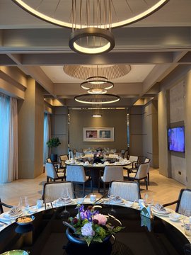The image shows a large dining room with multiple tables set up for guests, each table adorned with various items such as wine glasses, cups, bowls, and vases. The chairs surrounding the tables are arranged neatly, creating an inviting atmosphere for socializing or dining. A potted plant is placed on one of the tables, adding a touch of greenery to the room. In addition to these elements, there are several wine glasses scattered around the space, suggesting that guests may be enjoying drinks during their visit. The overall scene conveys a sense of elegance and sophistication, with attention given to both the interior design and the presentation of food and beverages for the event.
