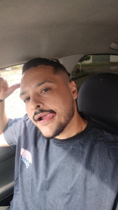 Selfie of a young man with a beard sitting in a car, sticking his tongue out and saluting, wearing a dark blue t-shirt.