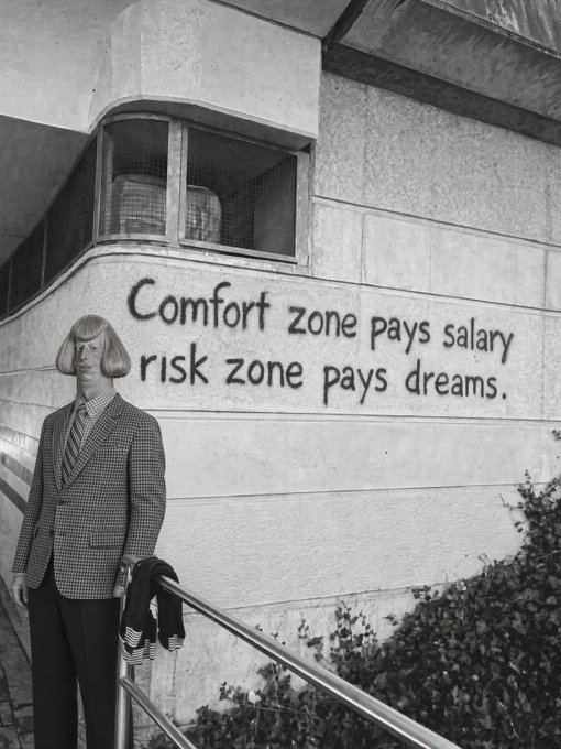 A black and white photo of a man in a checkered blazer and tie standing on steps, with the text on the wall behind him reading 'Comfort zone pays salary, risk zone pays dreams.'