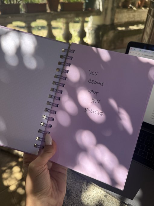 A hand holds an open purple spiral notebook with the handwritten message 'YOU BECOME WHAT YOU BELIEVE', illuminated by sunlight filtering through leaves, with a laptop visible in the background.