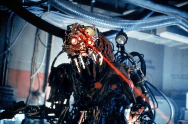 A menacing cyborg-like creature with glowing red eyes and a red laser beam shooting from its mouth in a dimly lit industrial setting.