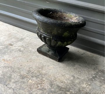 A weathered, dark stone urn or planter with classical detailing sits on a concrete surface in front of gray siding.