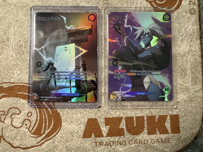 Two Azuki Trading Card Game cards displayed on a playmat: 'Surge Gate' and 'Raizan', both featuring α (alpha) designation with holographic/foil finishes.
