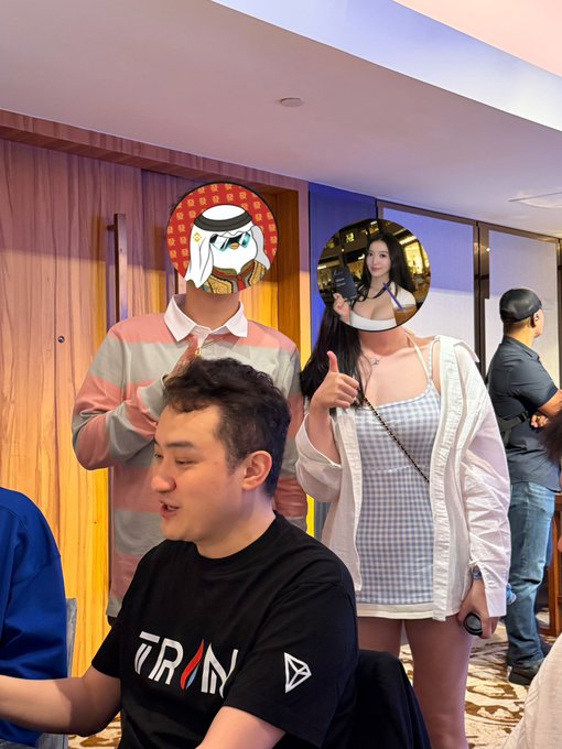A group of people pose indoors, with two faces obscured by circular sticker images, one resembling a cartoon character in a headdress.