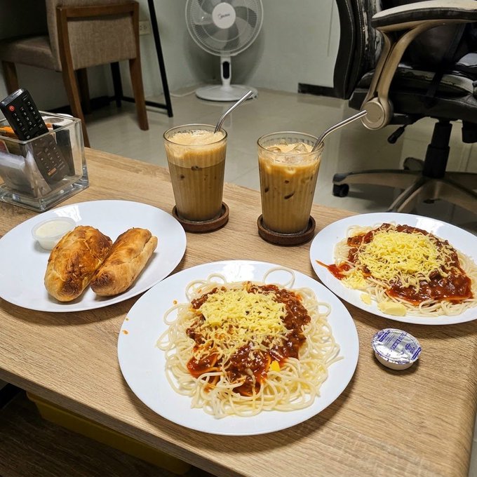 A meal is spread on a wooden table featuring two glasses of iced coffee, two plates of spaghetti topped with red sauce and shredded cheese, a plate with two pastries, a small butter dish, and a yogurt container, set in what appears to be an office space with a fan and chairs in the background.