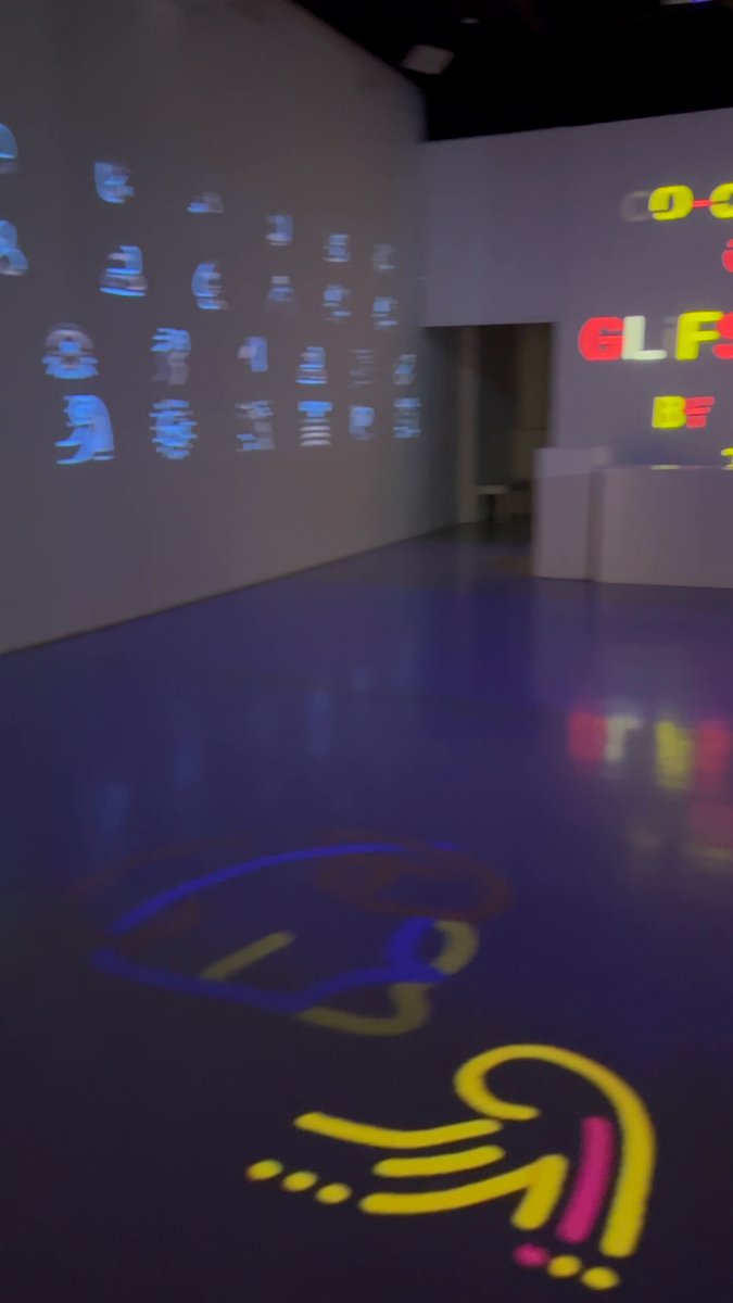 The image shows an art installation featuring a large projection of colorful letters and shapes on a wall, which appears to be illuminated by blue light. The text "Standing in the room at Lume watching your GLiFS go up. I’ve been making art a long time. I’ve never gotten to see a whole community's work breathing on the walls at once like this." is displayed along with the image, and there are also some words that read "partiful" and "RSVP". The scene seems to be set in an art gallery or exhibition space where people can come and experience the unique artwork.