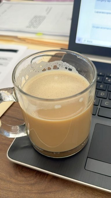 A glass mug filled with a light brown coffee drink with foam on top, sitting on the keyboard of a laptop with a webpage open in the background.
