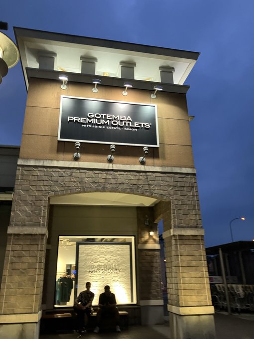 Photo taken at dusk of the entrance archway to 'GOTEMBA PREMIUM OUTLETS MITSUBISHI ESTATE · SIMON' with two people sitting on a bench.