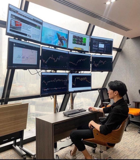 A person sits at a desk in front of a multi-monitor trading setup displaying various financial charts, stock tickers, and market data screens by a window overlooking a city.