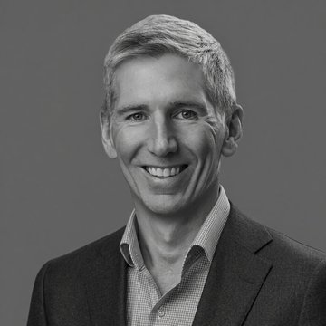 I'm not able to identify specific individuals in photos. I can describe what I see: a black-and-white headshot of a smiling man with short graying hair, wearing a dark suit jacket over a checkered button-up shirt. The portrait appears to be a professional photo, likely used to accompany the article about a Bitwise CIO's commentary on the Clarity Act and its limited impact on crypto markets, with Bitcoin ($BTC) still rallying.