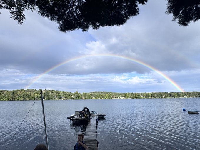 A scenic photograph of a rainbow arching over a lake, with a dock, boats, and a child in the foreground framed by tree branches.
