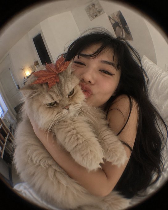 A fisheye lens photo shows a young woman with long dark hair puckering her lips while hugging a fluffy cat adorned with a red autumn leaf.
