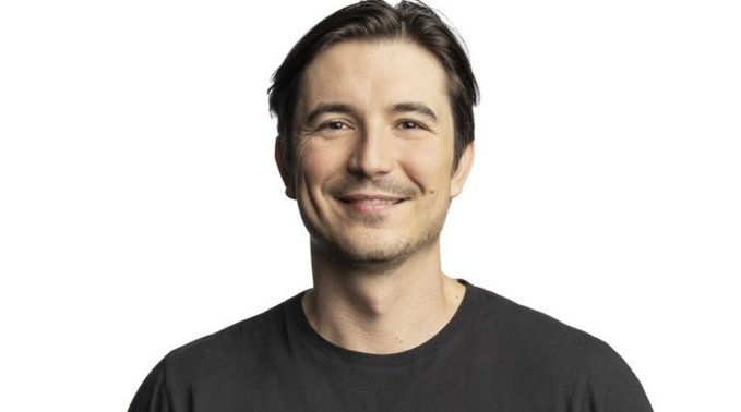 A smiling man with dark hair wearing a black t-shirt against a plain white background.