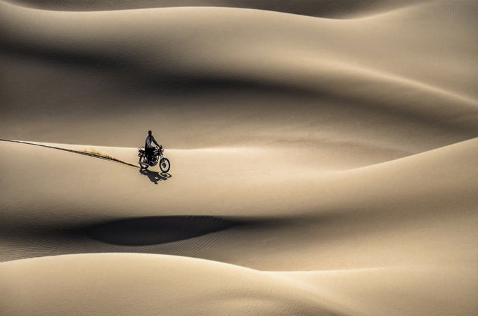 An aerial view of a person riding a motorcycle across expansive, rippling sand dunes, casting a long shadow on the tan sand.