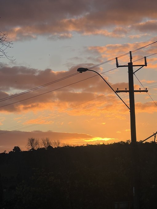 A sunset scene with orange and pink clouds, featuring silhouetted power lines, a utility pole, and a street lamp against the twilight sky.