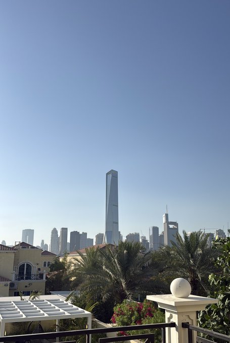 A view of a modern city skyline featuring a tall, sleek skyscraper, likely the Zifeng Tower in Nanjing, framed by palm trees and a villa under a clear blue sky.