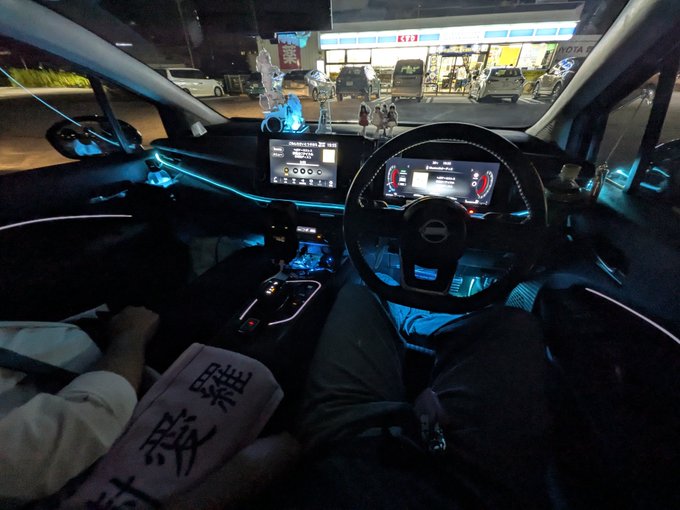 View from the passenger seat of a car at night, showing a driver, illuminated dashboard and steering wheel, with a brightly lit 'E&S' convenience store visible through the windshield.