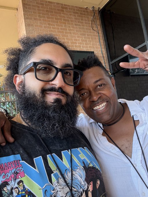 Two men pose for a cheerful selfie outdoors, with one wearing glasses, a beard, and a black 'MARVEL' X-Men t-shirt, while the other smiles broadly in a white shirt.