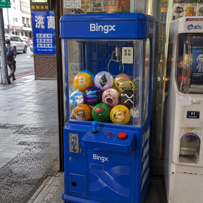 This BingX claw machine is filled with cryptocurrency-themed balls, like Bitcoin, Ethereum, Solana, Dogecoin, and Shiba the Inu, inviting players to try their luck. The playful nature of the claw machine aligns perfectly with the post's invitation to "Grab it!" and choose a prize.
