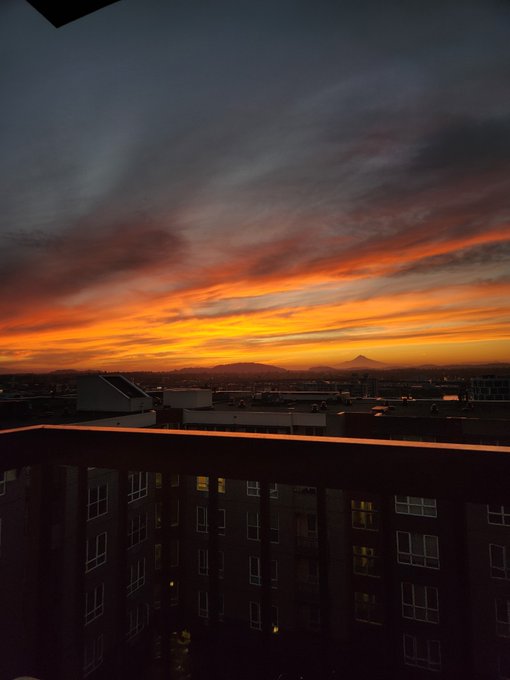 A vibrant sunset with orange and pink clouds over a cityscape viewed from a balcony, with mountains visible in the distance.