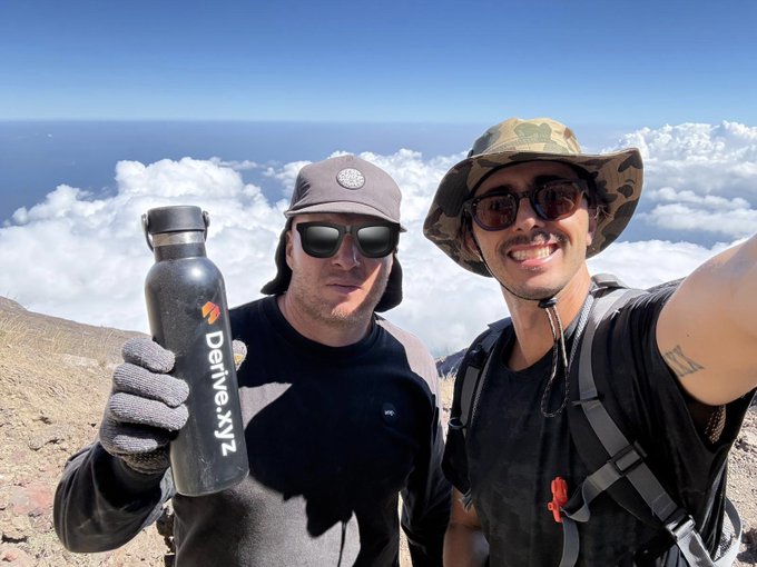 Two hikers take a selfie on a mountain summit above the clouds, one holding a black water bottle labeled 'Derive.xyz'.